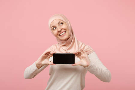 Pensive young arabian muslim woman in hijab light clothes posing isolated on pink background. People...