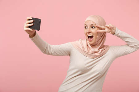 Excited arabian muslim woman in hijab light clothes posing isolated on pink background. People religious lifestyle concept. Mock up copy space. Doing selfie shot on mobile phone, showing victory sign.の写真素材