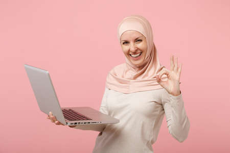 Joyful young arabian muslim woman in hijab light clothes posing isolated on pink background. People religious lifestyle concept. Mock up copy space. Hold, work laptop pc computer, showing OK gesture.の写真素材