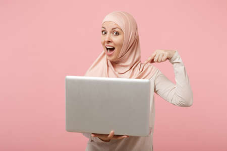 Excited young arabian muslim woman in hijab light clothes posing isolated on pink background. People religious Islam lifestyle concept. Mock up copy space. Pointing index finger on laptop pc computer.の写真素材