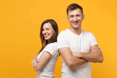 Cheerful young couple two friends guy girls in white blank empty design t-shirts posing isolated on yellow orange wall background. People lifestyle concept. Mock up copy space. Holding hands crossed.の写真素材