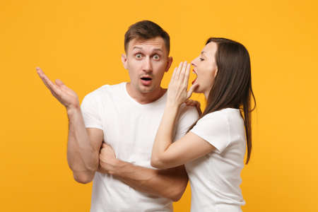Young couple two friends guy girls in white blank empty design t-shirts posing isolated on yellow orange wall background. People lifestyle concept. Mock up copy space. Whispering secret behind hand.の写真素材