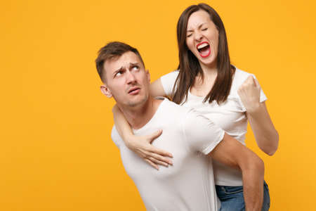 Young couple in white t-shirts posing isolated on yellow orange background. People lifestyle concept. Mock up copy space. Giving piggyback ride to joyful sitting on back, clenching fist like winner.の写真素材