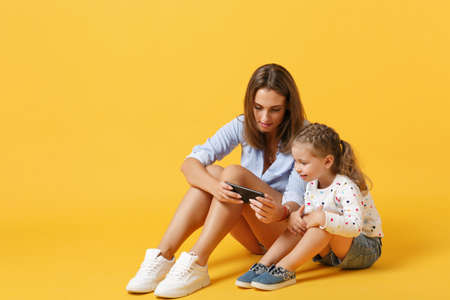 Woman using mobile phone have fun with cute child baby girl 4-5 years old. Mommy little kid daughter isolated on yellow background studio portrait Mothers Day love family parenthood childhood conceptの写真素材