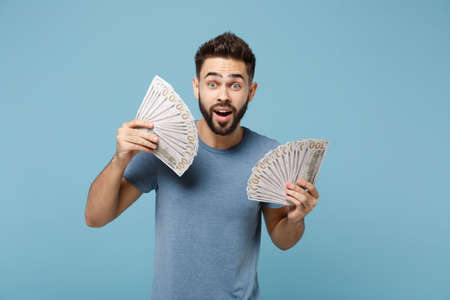 Young surprised man in casual clothes posing isolated on blue background, studio portrait. People sincere emotions lifestyle concept. Mock up copy space. Holding fan of cash money in dollar banknotes.の写真素材