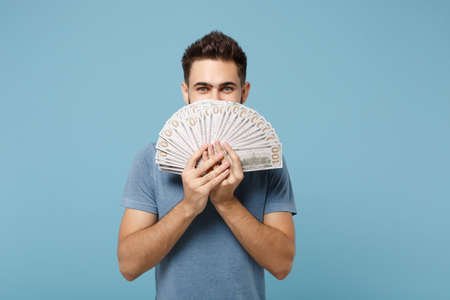 Young handsome man in casual clothes posing isolated on blue wall background, studio portrait. People lifestyle concept. Mock up copy space. Covering face with fan of cash money in dollar banknotes.の写真素材