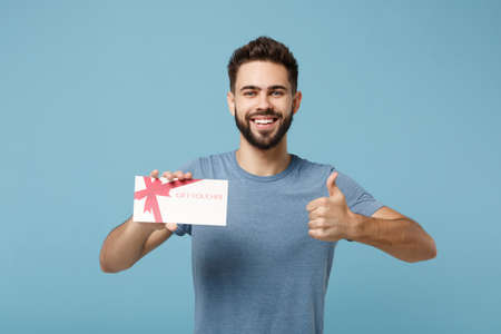 Young smiling man in casual clothes posing isolated on blue wall background, studio portrait. People sincere emotions lifestyle concept. Mock up copy space. Holding gift certificate, showing thumb up.