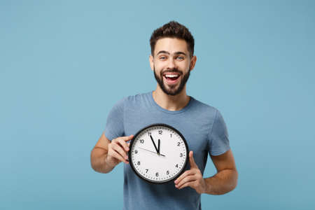 Young cheerful laughing man in casual clothes posing isolated on blue wall background, studio portrait. People sincere emotions lifestyle concept. Mock up copy space. Holding in hands round clock.の写真素材