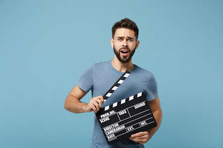 Young shocked man in casual clothes posing isolated on blue wall background, studio portrait. People lifestyle concept. Mock up copy space. Holding in hands classic black film making clapperboard.の写真素材