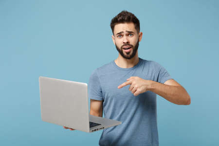 Young perplexed confused man in casual clothes posing isolated on blue wall background, studio portrait. People lifestyle concept. Mock up copy space. Pointing index finger on laptop pc computer.の写真素材