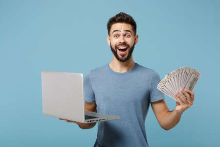 Young excited surprised man in casual clothes posing isolated on blue wall background. People lifestyle concept. Mock up copy space. Holding laptop pc computer, fan of cash money in dollar banknotes.の写真素材