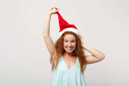 Smiling young redhead Santa girl in light clothes posing isolated on white wall background, studio portrait. Happy New Year 2020 celebration holiday concept. Mock up copy space. Holding Christmas hat.の写真素材
