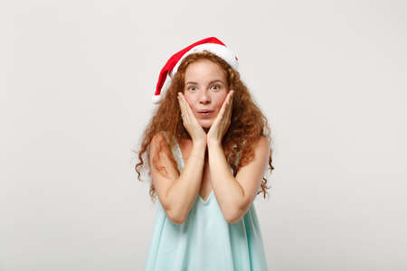 Amazed young redhead Santa girl in light clothes, Christmas hat isolated on white background in studio. Happy New Year 2020 celebration holiday concept. Mock up copy space. Putting hands on cheeks.の写真素材