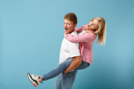 Young fun couple two friends guy girl in white pink empty blank design t-shirts posing isolated on pastel blue background studio portrait. People lifestyle concept. Mock up copy space. sitting on backの写真素材