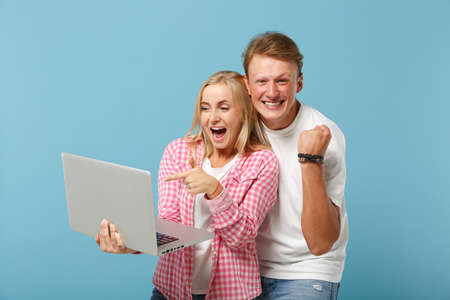 Young happy couple friends guy girl in white pink empty blank t-shirts posing isolated on pastel blue background. People lifestyle concept. Mock up copy space. Pointing finger on laptop pc computer.の写真素材