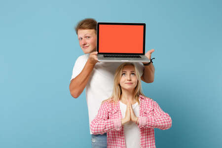 Young couple friends guy girl in white pink t-shirts posing isolated on blue background. People lifestyle concept. Mock up copy space. Hold laptop computer with empty screen, hands folded in prayer.の写真素材
