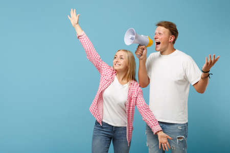 Young cheerful couple two friends guy girl in white pink empty t-shirts posing isolated on pastel blue background. People lifestyle concept. Mock up copy space. Scream in megaphone, spreading hands.の写真素材