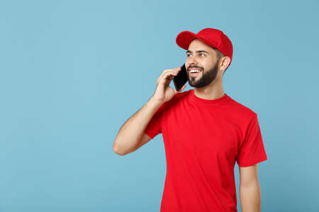 Delivery man in red uniform workwear hold cellphone isolated on blue background, studio portrait. Professional male employee in cap t-shirt working courier dealer. Service concept. Mock up copy space.の写真素材