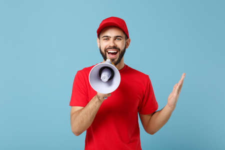 Delivery man in red workwear scream in megaphone isolated on blue background studio portrait....