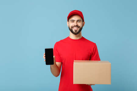Delivery man in red uniform isolated on blue background, studio portrait. Male employee in cap t-shirt print working as courier dealer hold empty cardboard box. Service concept. Mock up copy space.の写真素材