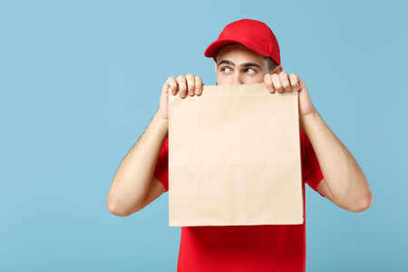 Delivery man in red uniform hold craft paper packet with food isolated on blue background, studio...