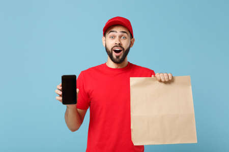Delivery man in red uniform hold craft paper packet with food isolated on blue background, studio...
