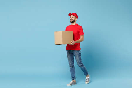 Delivery man in red uniform isolated on blue background, studio portrait. Male employee in cap t-shirt print working as courier dealer hold empty cardboard box. Service concept. Mock up copy space.の写真素材