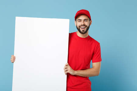 Delivery man in red workwear hold big white empty billboard isolated on blue background, studio...