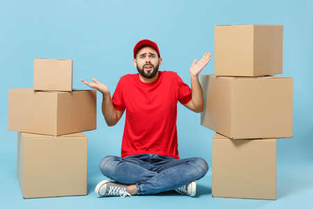 Delivery sad man in red uniform isolated on blue background, studio portrait. Male employee in cap t-shirt print working as courier dealer hold empty cardboard box. Service concept. Mock up copy spaceの写真素材
