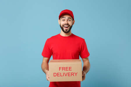 Delivery man in red uniform isolated on blue background, studio portrait. Male employee in cap t-shirt print working as courier dealer hold empty cardboard box. Service concept. Mock up copy space.の写真素材