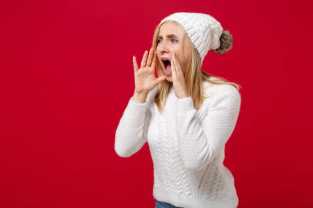Young woman in white sweater hat isolated on red background studio portrait. Healthy fashion lifestyle people emotions cold season concept. Mock up copy space. Screaming with hands gesture near mouth.の写真素材