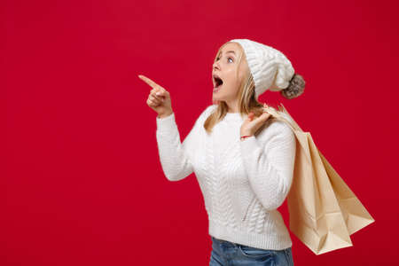 Amazed woman in sweater hat isolated on red background. Healthy fashion lifestyle, cold season concept. Mock up copy space. Hold package bag with purchases after shopping, pointing index finger aside.の写真素材