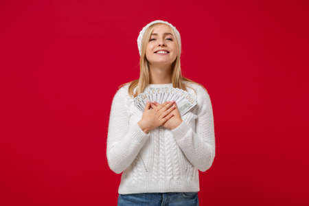 Smiling young woman in white sweater, hat isolated on red background, studio portrait. Healthy fashion lifestyle, cold season concept. Mock up copy space. Hold fan of cash money in dollar banknotes.の写真素材