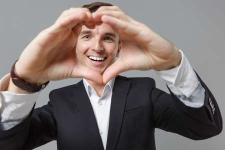 Smiling young business man in classic suit shirt posing isolated on grey background. Achievement career wealth business concept. Mock up copy space. Showing shape heart with hands, heart-shape sign.の写真素材