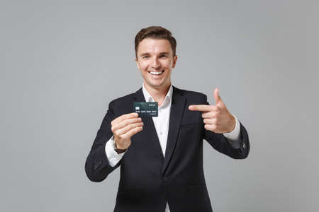 Funny young business man in classic black suit shirt posing isolated on grey wall background. Achievement career wealth business concept. Mock up copy space. Pointing index finger on credit bank card.の写真素材
