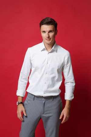 Attractive young business man in white shirt, gray pants posing isolated on bright red wall background studio portrait. Achievement career wealth business concept. Mock up copy space. Looking camera.の写真素材