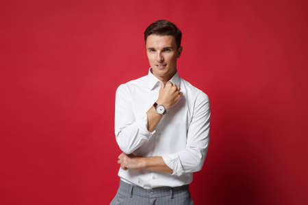 Handsome young business man in white shirt, gray pants posing isolated on bright red wall background in studio. Achievement career wealth business concept. Mock up copy space. Keeping hand on collar.の写真素材