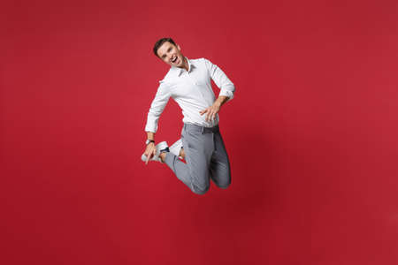 Cheerful funny young business man in white shirt, gray pants posing isolated on bright red wall background studio portrait. Achievement career wealth business concept. Mock up copy space. Jumping.の写真素材
