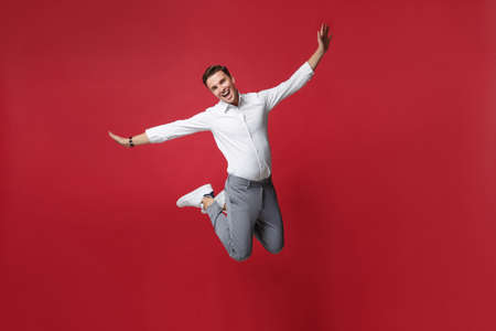 Funny young business man in white shirt, gray pants posing isolated on bright red background studio portrait. Achievement career wealth business concept. Mock up copy space. Jumping, spreading hands.の写真素材