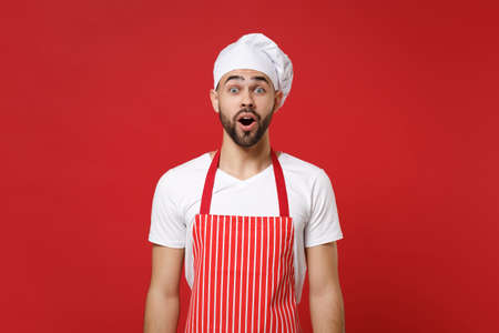 Shocked young bearded male chef cook or baker man in striped apron white t-shirt toque chefs hat posing isolated on red background. Cooking food concept. Mock up copy space. Keeping mouth wide open.の写真素材