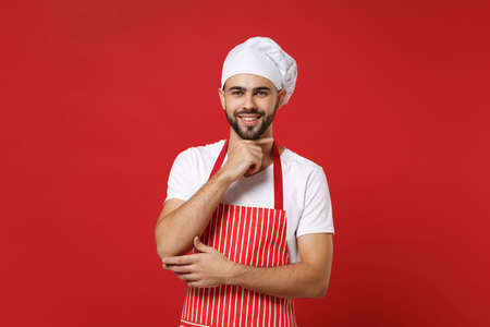 Smiling young bearded male chef cook or baker man in striped apron white t-shirt toque chefs hat posing isolated on red background. Cooking food concept. Mock up copy space. Put hand prop up on chin.の写真素材