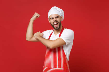 Strong young bearded male chef cook or baker man in striped apron white t-shirt toque chefs hat posing isolated on red background. Cooking food concept. Mock up copy space. Showing biceps, muscles.の写真素材