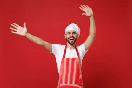 Joyful bearded male chef cook or baker man in striped apron t-shirt toque chefs hat posing isolated on red background. Cooking food concept. Mock up copy space. Expressive gesticulating with hands.の写真素材