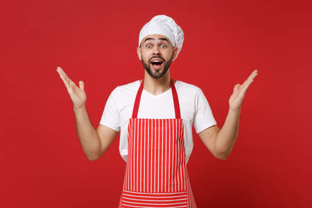 Shocked bearded male chef cook or baker man in striped apron t-shirt toque chefs hat posing isolated on red background. Cooking food concept. Mock up copy space. Keeping mouth open, spreading hands.の写真素材
