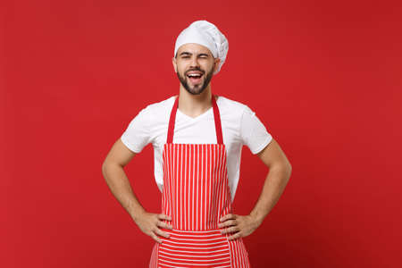 Funny bearded male chef cook or baker man in striped apron white t-shirt toque chefs hat posing isolated on red background. Cooking food concept. Mock up copy space. Blinking standing with arm akimbo.の写真素材
