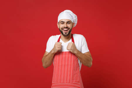 Joyful young bearded male chef cook or baker man in striped apron white t-shirt toque chefs hat posing isolated on red background in studio. Cooking food concept. Mock up copy space. Clenching fists.の写真素材