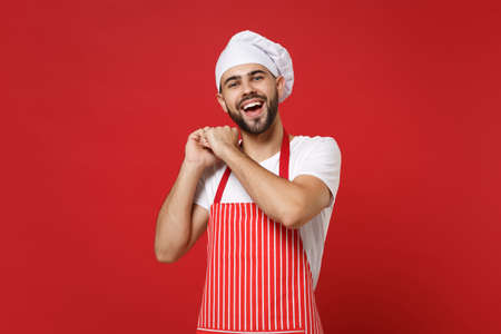 Funny young bearded male chef cook or baker man in striped apron white t-shirt toque chefs hat posing isolated on red background in studio. Cooking food concept. Mock up copy space. Clenching fists.の写真素材