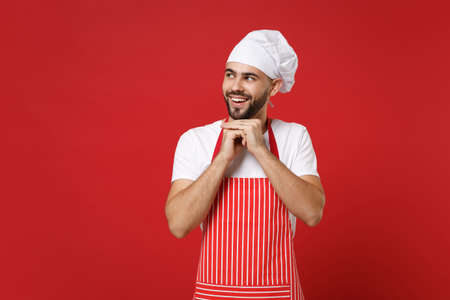Smiling young bearded male chef cook or baker man in striped apron toque chefs hat posing isolated on red background. Cooking food concept. Mock up copy space. Looking aside put hands prop up on chin.の写真素材