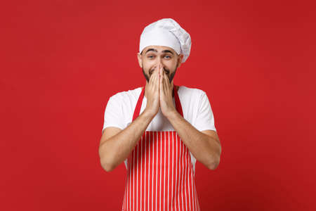 Excited young bearded male chef cook or baker man in striped apron white t-shirt toque chefs hat posing isolated on red background. Cooking food concept. Mock up copy space. Covering mouth with hands.の写真素材