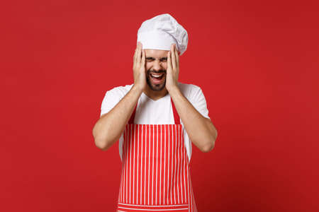 Screaming bearded male chef cook or baker man in striped apron toque chefs hat posing isolated on red background. Cooking food concept. Mock up copy space. Keeping eyes closed putting hands on head.の写真素材